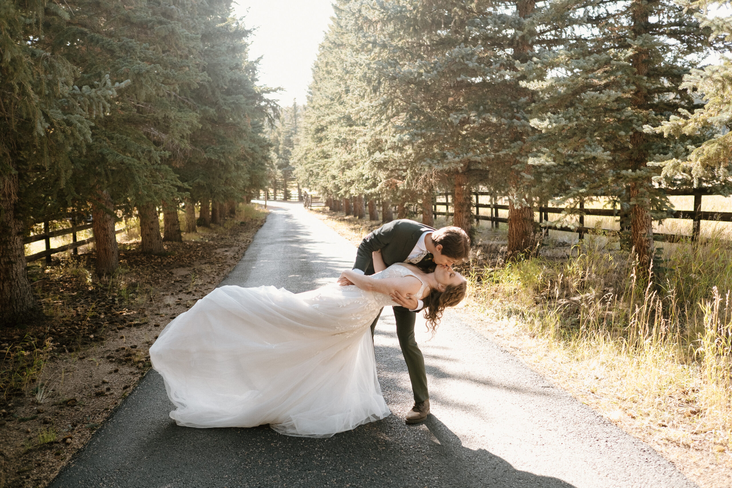 Groom dips bride and they share a kiss on a road surrounded by pine trees in Colorado